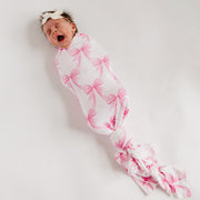 A sleeping newborn baby wrapped in a white swaddle blanket with a pink bow pattern, wearing a matching white headband with a pink bow, photographed on a plain light background.