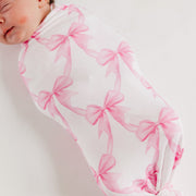 A sleeping newborn baby wrapped in a soft white swaddle blanket featuring a repeating pink bow pattern, photographed against a clean white background.