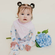 A baby girl sitting on a cream studio background wearing a light pink floral bib over a white bunny-printed long-sleeve romper, with a bouquet of blue hydrangeas next to her.