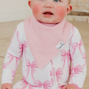 A close-up of a baby wearing a white long-sleeve bodysuit with a pink ribbon bow pattern and a matching pink bib. The bodysuit has a white zipper down the front.