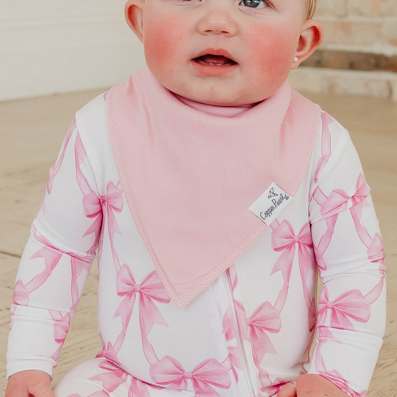 A close-up of a baby wearing a white long-sleeve bodysuit with a pink ribbon bow pattern and a matching pink bib. The bodysuit has a white zipper down the front.