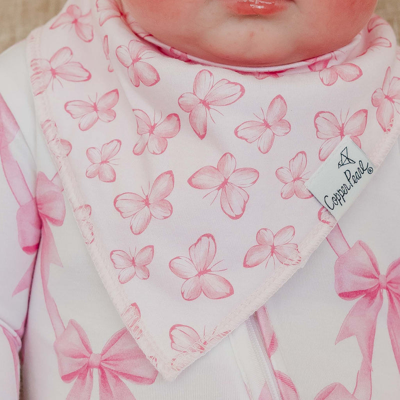 Close-up of a baby wearing a white bib with a pink butterfly and bow pattern, attached to a white bodysuit. A brand tag reading 'Copper Paws' is visible on the bib.