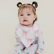 A baby wearing a white long-sleeve footed sleeper with pink bow and blue floral prints, along with a white bandana bib featuring pink bows, set against a plain light beige background.