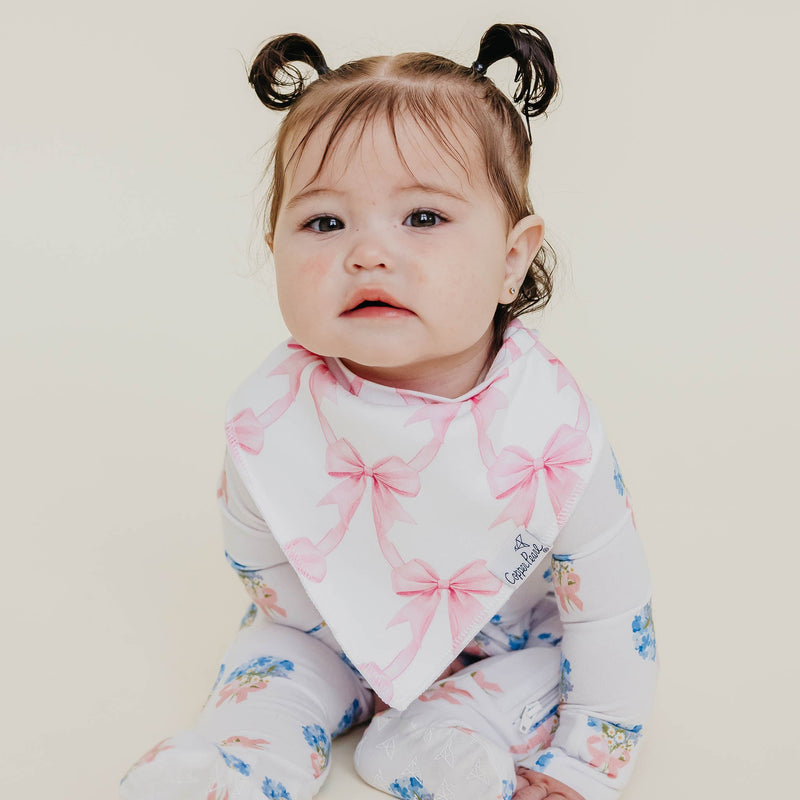 A baby wearing a white long-sleeve footed sleeper with pink bow and blue floral prints, along with a white bandana bib featuring pink bows, set against a plain light beige background.