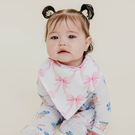 A baby wearing a white long-sleeve footed sleeper with pink bow and blue floral prints, along with a white bandana bib featuring pink bows, set against a plain light beige background.