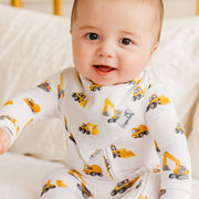 A baby wearing a white long-sleeve footless onesie with a yellow and gray construction vehicle print, sitting on a white quilted bed.