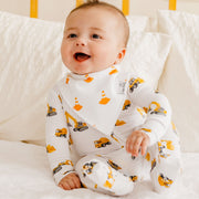 A baby sitting on a white bed wearing a white footed sleeper with a yellow and orange construction print and a matching bib.