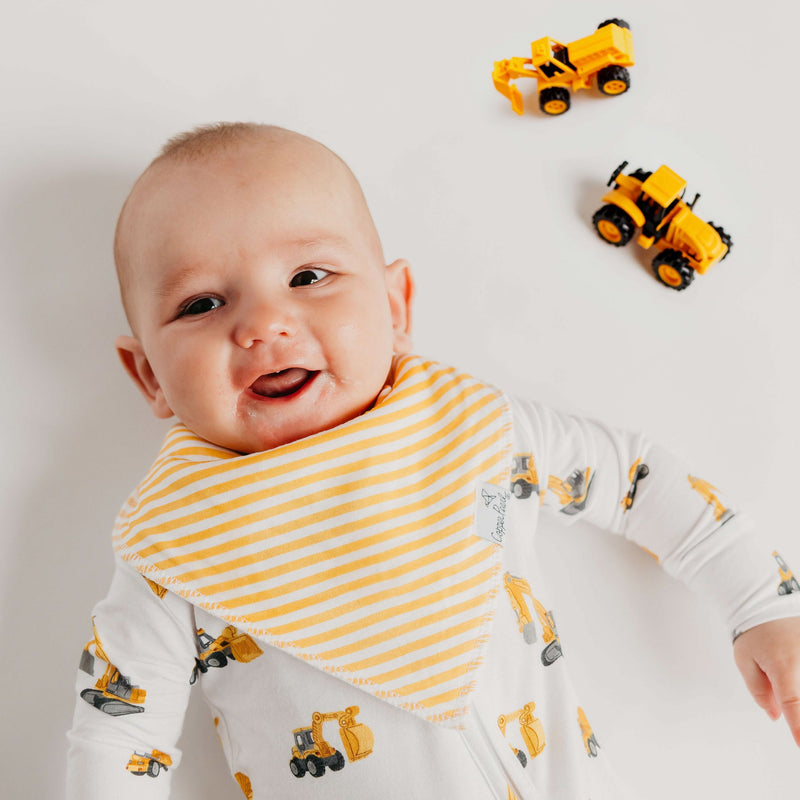 A smiling baby wearing a yellow and white striped bib over a white long-sleeve romper with yellow construction vehicle prints, lying on a white background with two toy construction vehicles nearby.