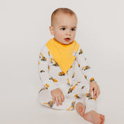 A baby sitting on a light gray studio background wearing a white long-sleeve footless jumpsuit with a yellow construction vehicle print and a solid yellow bib.