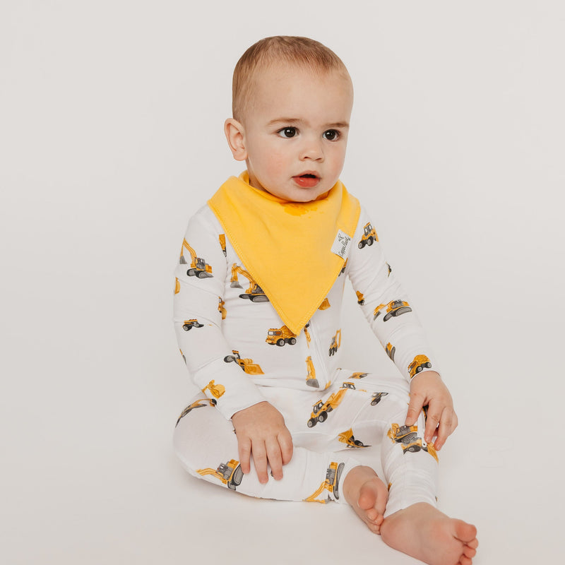 A baby sitting on a light gray studio background wearing a white long-sleeve footless jumpsuit with a yellow construction vehicle print and a solid yellow bib.