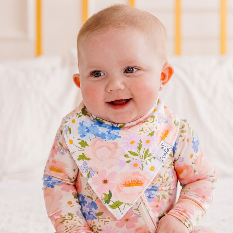 A smiling baby wearing a pink floral long-sleeve bodysuit and a matching floral bib, sitting on a white quilted surface.