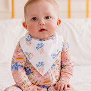 A baby wearing a pink floral long-sleeve sleep suit and a white bib with blue and pink floral prints, sitting on a white bed.