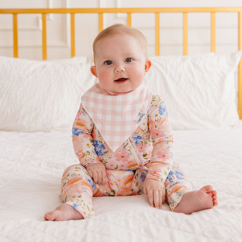 A baby sitting on a white quilted bed wearing a floral long-sleeve romper with a pink and white gingham bib. The background shows a yellow wooden crib headboard and white pillows.