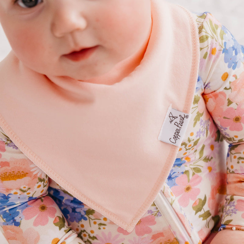 A close-up of a baby wearing a soft peach-pink triangular bib over a floral long-sleeve romper. A small white fabric tag on the bib reads 'Coconut Baby'.