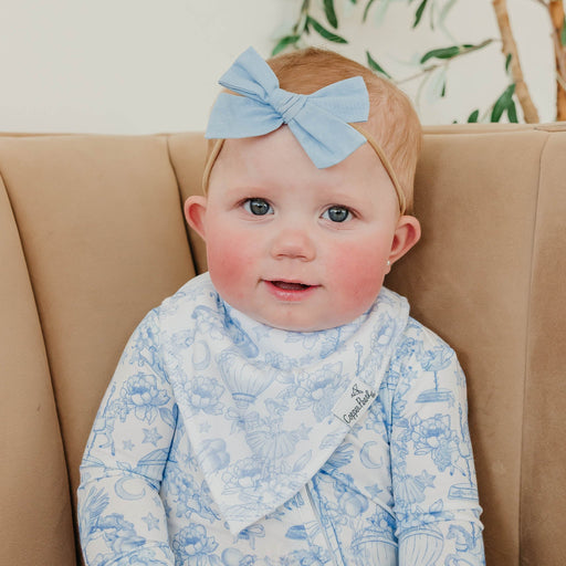 A baby wearing a light blue patterned bib and matching headband with a large bow, sitting on a beige armchair.