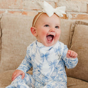 A baby wearing a blue and white toile-print one-piece jumpsuit and a matching bow headband, sitting on a beige couch.