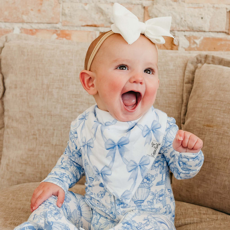 A baby wearing a blue and white toile-print one-piece jumpsuit and a matching bow headband, sitting on a beige couch.