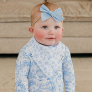 A baby wearing a light blue bow headband and a light blue patterned bib over a matching long-sleeve top, standing in front of a beige couch.