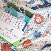 A stack of copper pearl premium burp cloths with a baseball theme, featuring pinstripes, red and blue baseballs, and glove prints, with a paper label wrapped around the front.