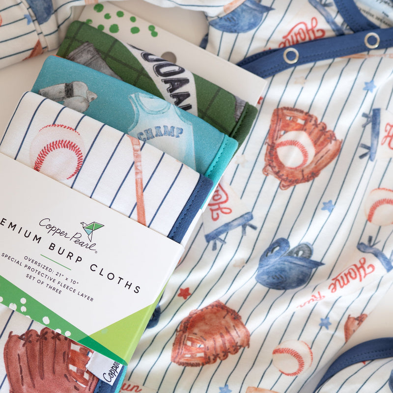 A stack of copper pearl premium burp cloths with a baseball theme, featuring pinstripes, red and blue baseballs, and glove prints, with a paper label wrapped around the front.
