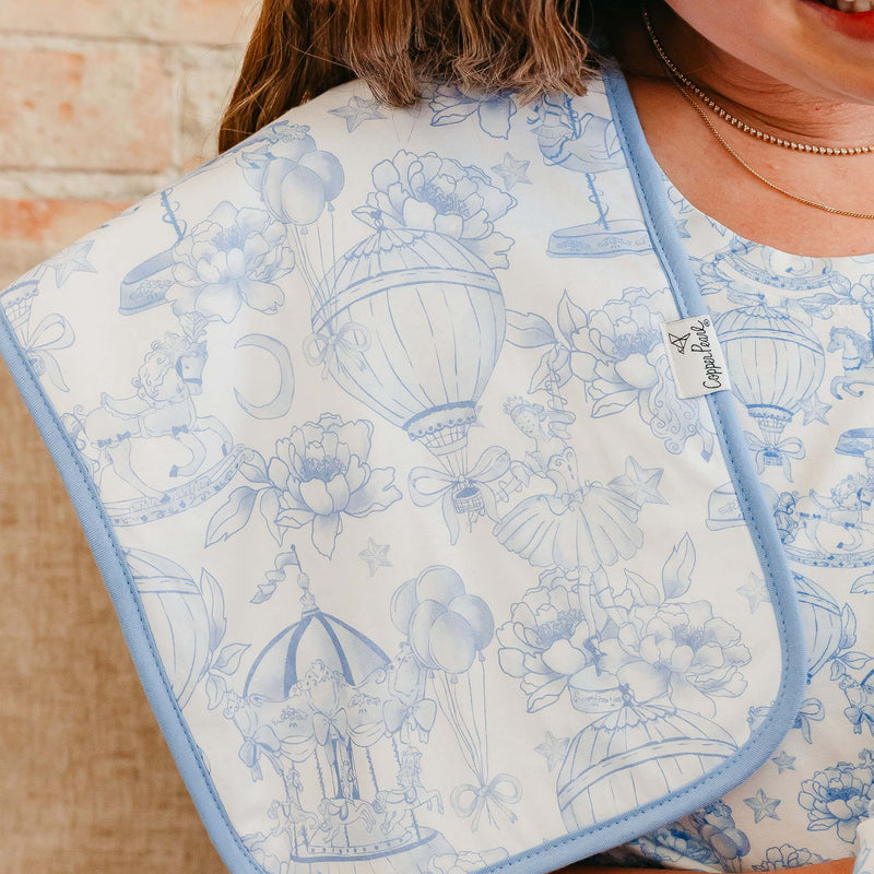 A close-up of a baby bib with a light blue and white toile-style print featuring hot air balloons, flowers, and teacups, worn by a person in front of a brick wall.