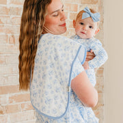 A woman wearing a light blue and white floral baby carrier wrap holds a baby wearing a matching long-sleeve outfit and a light blue bow headband, set against a light brick wall background.