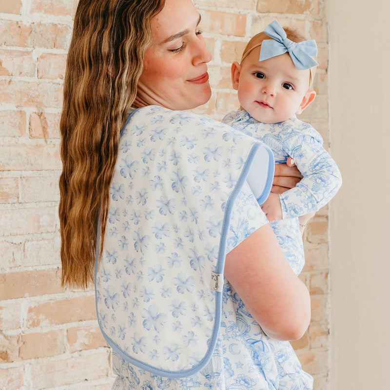 A woman wearing a light blue and white floral baby carrier wrap holds a baby wearing a matching long-sleeve outfit and a light blue bow headband, set against a light brick wall background.