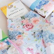 A styled flat-lay of Copper Pearl fabric accessories, including a packaged Bandana Bib and a Swaddle Blanket, both featuring a pastel floral print with pink, blue, and white flowers on a light blue background.