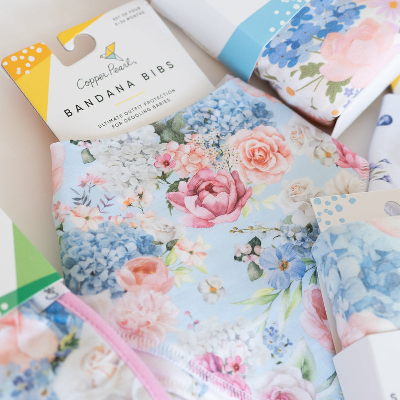 A styled flat-lay of Copper Pearl fabric accessories, including a packaged Bandana Bib and a Swaddle Blanket, both featuring a pastel floral print with pink, blue, and white flowers on a light blue background.