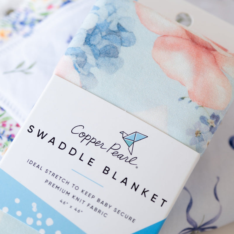 A packaged swaddle blanket from Copper Pearl, featuring a soft watercolor floral print in blue and pink on a white label, with other floral blankets blurred in the background.