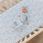 A baby lying on a fitted crib mattress wearing a matching light blue and white patterned one-piece outfit and headband.