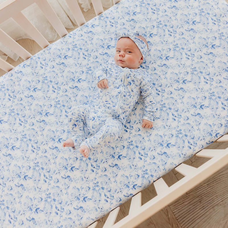 A baby lying on a fitted crib mattress wearing a matching light blue and white patterned one-piece outfit and headband.