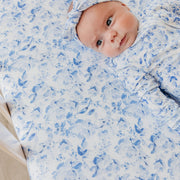 A baby lying on a white mattress or changing pad, wrapped in a light blue and white floral-patterned swaddle blanket and matching bonnet.