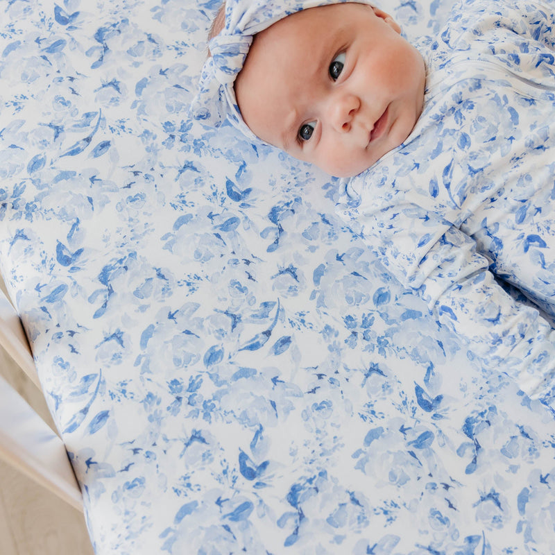A baby lying on a white mattress or changing pad, wrapped in a light blue and white floral-patterned swaddle blanket and matching bonnet.