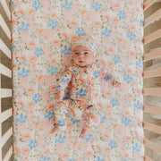 A baby lying on a floral crib mattress, wearing a matching floral sleeper and bonnet. The crib has light wood slats and a soft pink bedding setup.