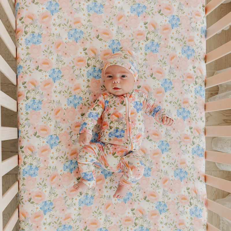 A baby lying on a floral crib mattress, wearing a matching floral sleeper and bonnet. The crib has light wood slats and a soft pink bedding setup.
