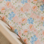 A close-up of a floral-patterned fabric, likely a crib bumper or mattress cover, draped over the corner of a light-colored wooden crib. The fabric features a delicate print of pink roses, blue hydrangeas, and small white daisies with green leaves on a cream background.