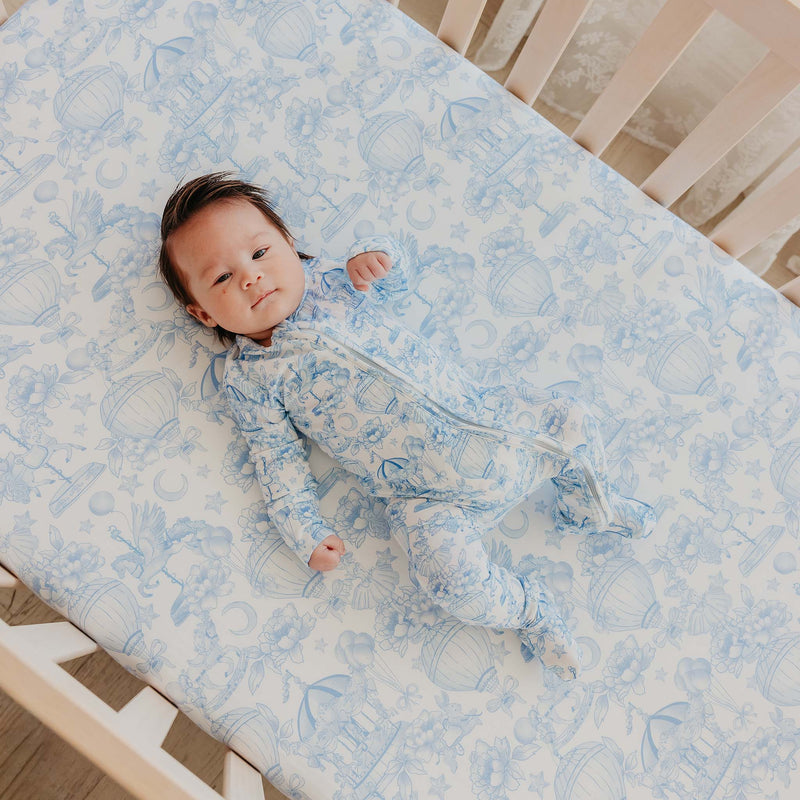 A baby lying on their back on a light blue and white patterned fitted sheet in a wooden crib, wearing a matching light blue and white patterned footed sleeper.