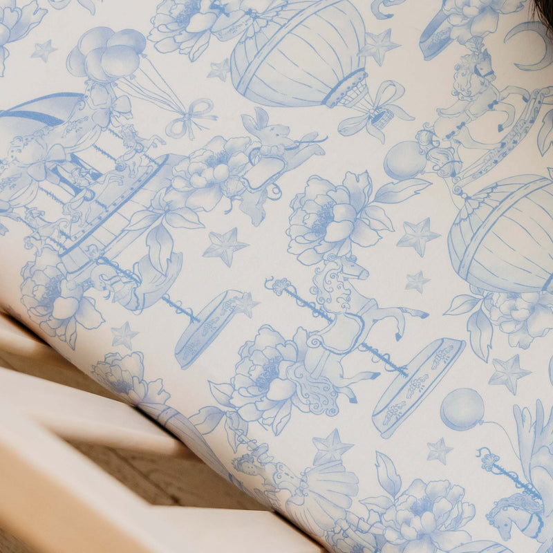 A close-up of a white wallpaper or fabric featuring a delicate blue toile-style pattern with hot air balloons, flowers, stars, and whimsical illustrations.