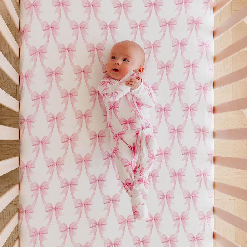 A baby wearing a white footed sleeper with a pink bow pattern lies on a matching crib mattress with a pink and white bow print.