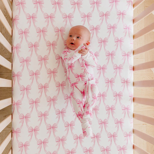 A baby wearing a white footed sleeper with a pink bow pattern lies on a matching crib mattress with a pink and white bow print.