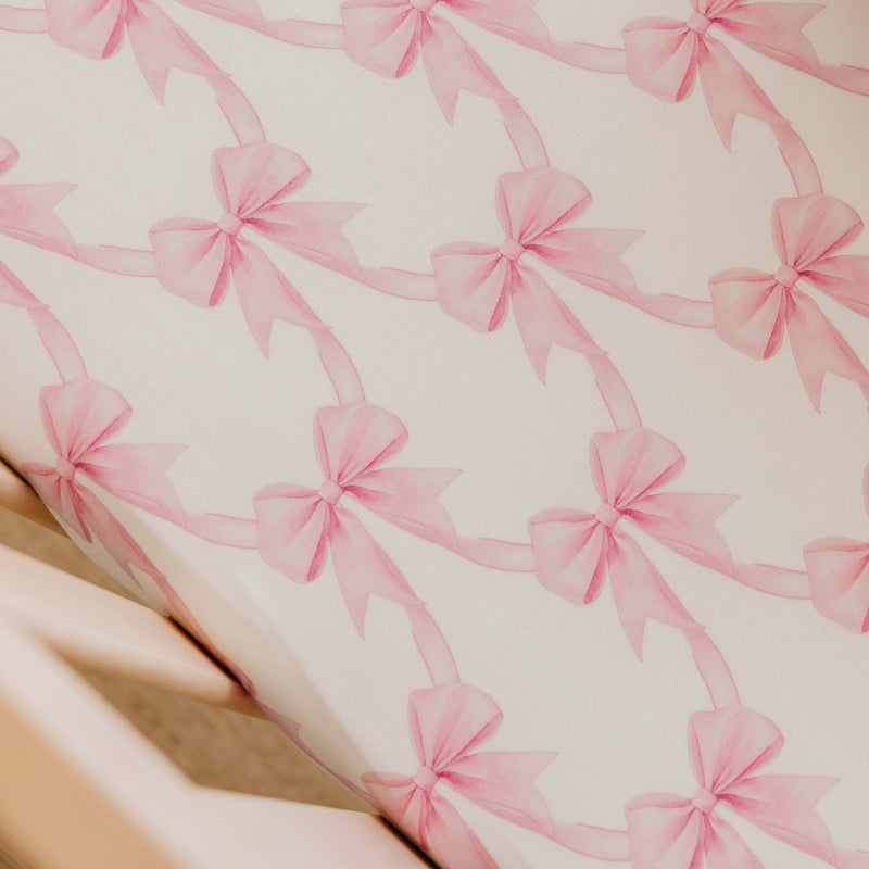 A close-up of a white or off-white fabric surface printed with a repeating pattern of soft pink ribbon bows connected by thin pink lines.