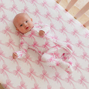 A baby lying on a crib mattress wearing a white footed sleeper with a pink bow pattern, next to matching crib bedding.
