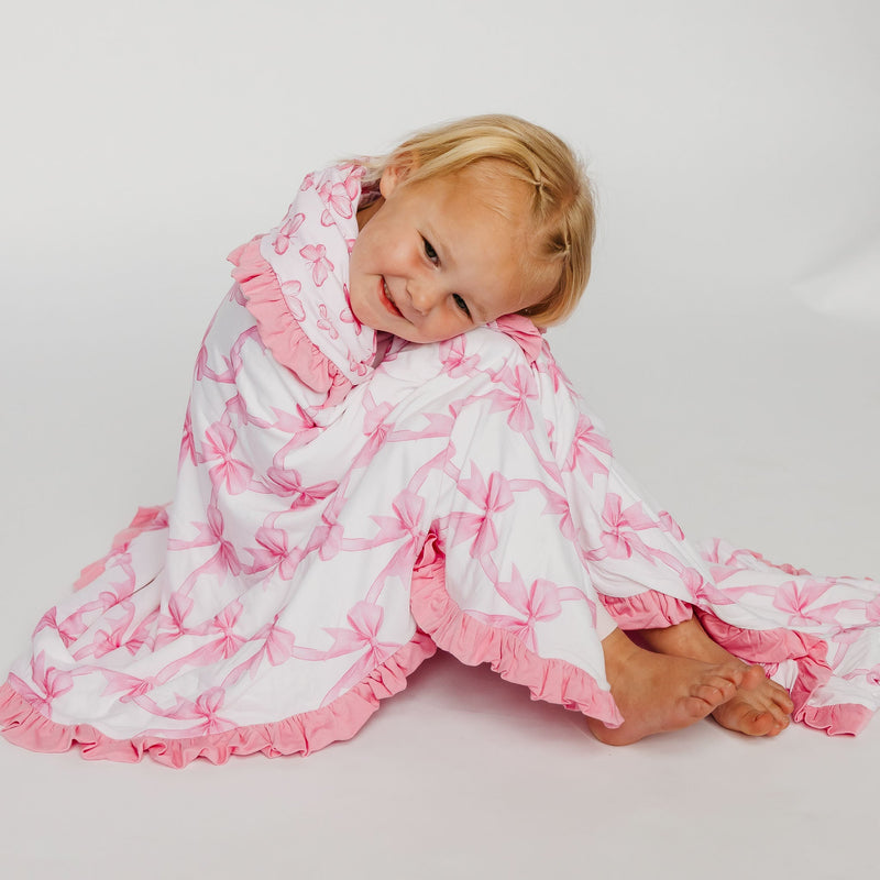 A smiling young child wrapped in a white cotton blanket with pink floral print and ruffled edges, posed on a light gray studio background.