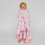 A toddler standing barefoot and wrapped in a white blanket with pink bow patterns and ruffled edges, photographed against a light gray studio background.
