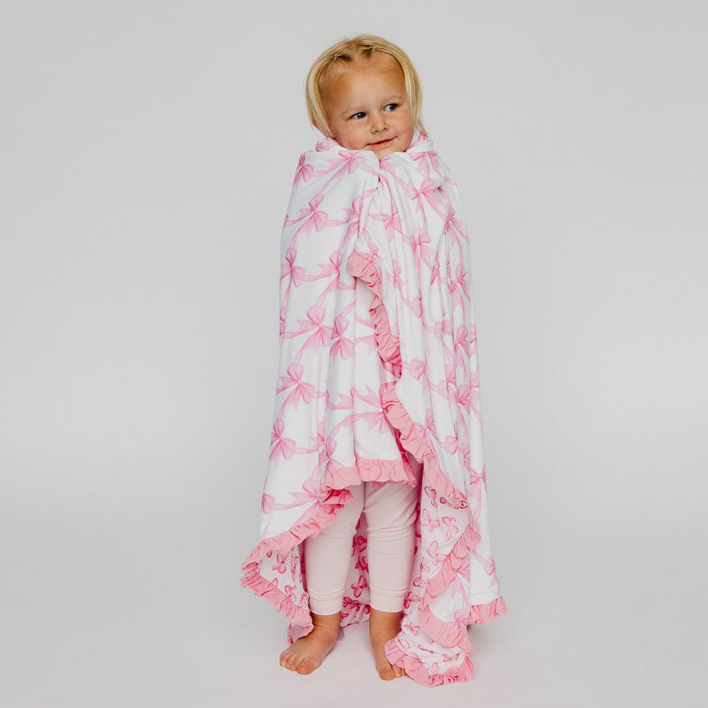 A toddler standing barefoot and wrapped in a white blanket with pink bow patterns and ruffled edges, photographed against a light gray studio background.