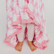 A child wearing light pink leggings stands barefoot under a white blanket with a pink bow pattern and ruffled edges, photographed against a plain light background.