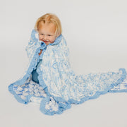 A smiling toddler wrapped in a light blue and white patterned blanket with ruffled edges, posed against a plain light gray studio background.