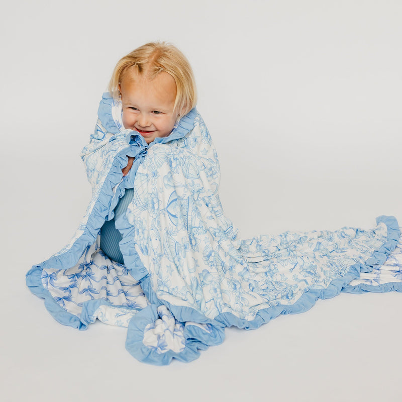 A smiling toddler wrapped in a light blue and white patterned blanket with ruffled edges, posed against a plain light gray studio background.
