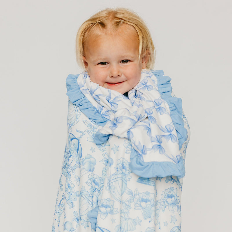 A young child with blonde hair is wrapped in a white and light blue patterned blanket with ruffled edges, photographed against a plain light gray background.